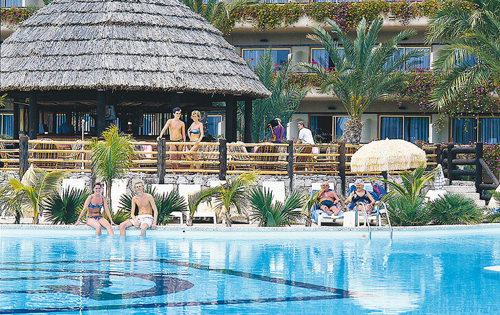 Hotelpool mit Palmen, Strohdach und Menschen in Badekleidung.