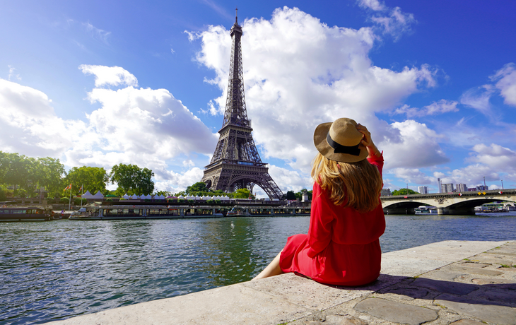 Reisende in rotem Kleid und Hut sitzt auf dem Kai der Seine und blickt auf den Eiffelturm