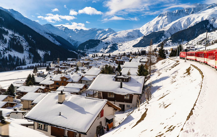 Glacier Express fährt durch eine verschneite Landschaft oberhalb eines sonnigen Dorfes