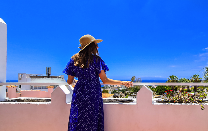 Rückansicht einer Touristin mit Sommerhut und blauem Kleid auf einem Balkon eines Luxusresort-Hotels in Costa Adeje, Teneriffa