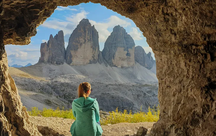 eine Frau sitzt vor einer Höhle, mit Blick auf die Tre Cime (Drei Zinnen) in den italienischen Dolomiten