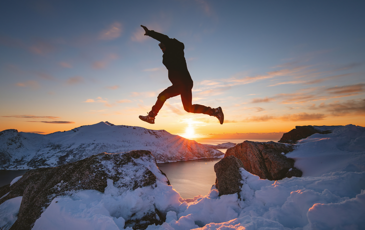 Silhouette einer Person, die auf Kvaloya in Norwegen zwischen schneebedeckten Felsen über einem Fjord hin und her springt.